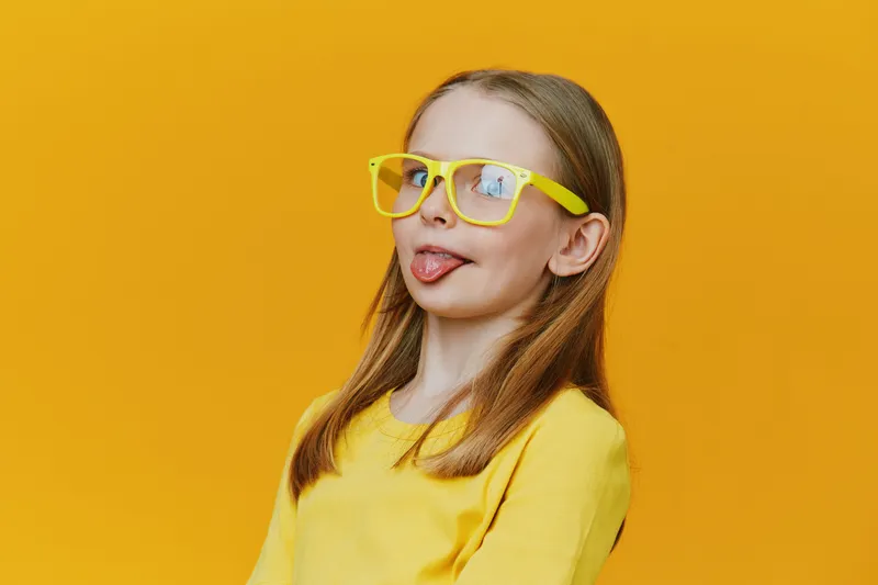 Playful girl with yellow glasses sticking out her tongue, showcasing a fun and energetic personality against a vibrant yellow background Bright colors evoke joy