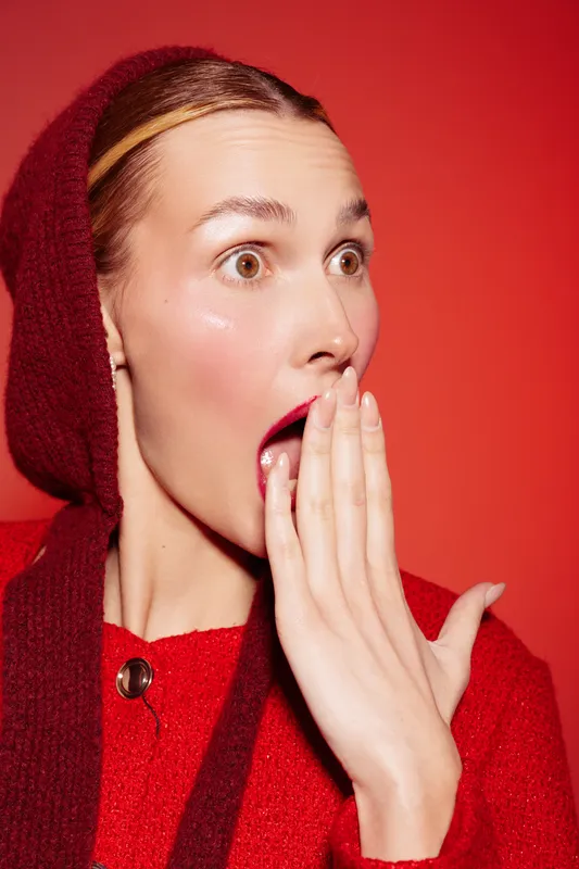Surprised woman with wide eyes and open mouth covers it with hand. She wears red sweater and hood against red background. Concept of shock, surprise, and unexpected emotions.