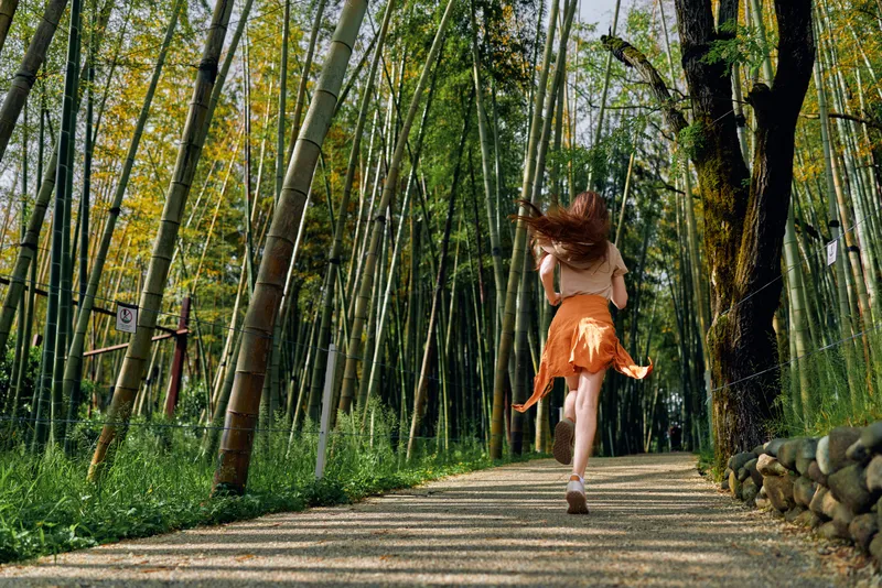 Woman running along a gravel path through a dense bamboo forest wearing an orange skirt, barefoot and in motion, back view framed by tall green trunks and summer sunlight.