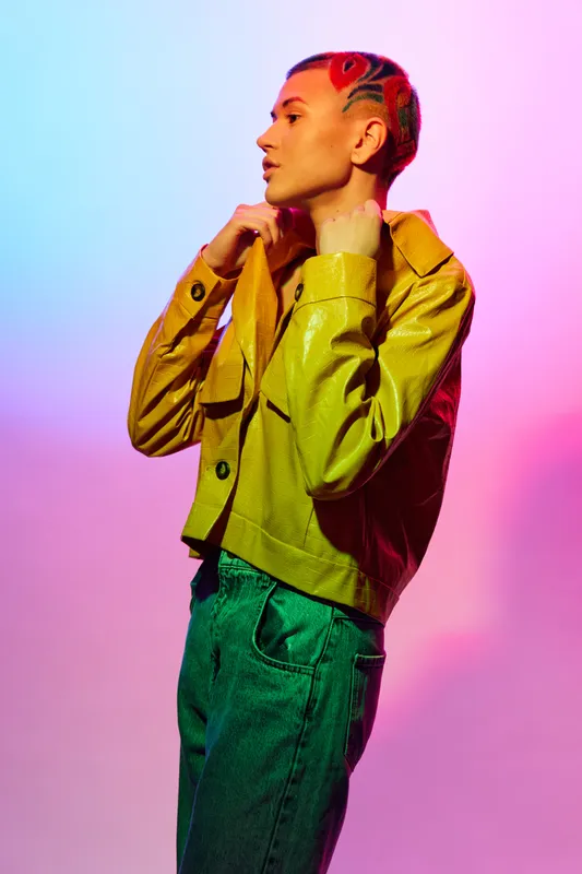 Woman with colorful hair and yellow jacket posing confidently against vibrant gradient background, showcasing modern fashion and artistic style.