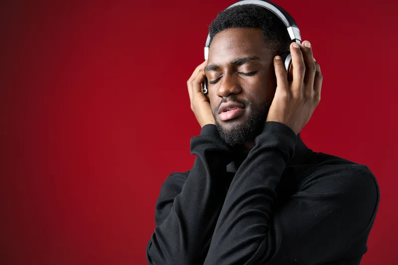 Young African American man enjoying music with headphones on a red background, expressing joy and relaxation through a closed eye smile