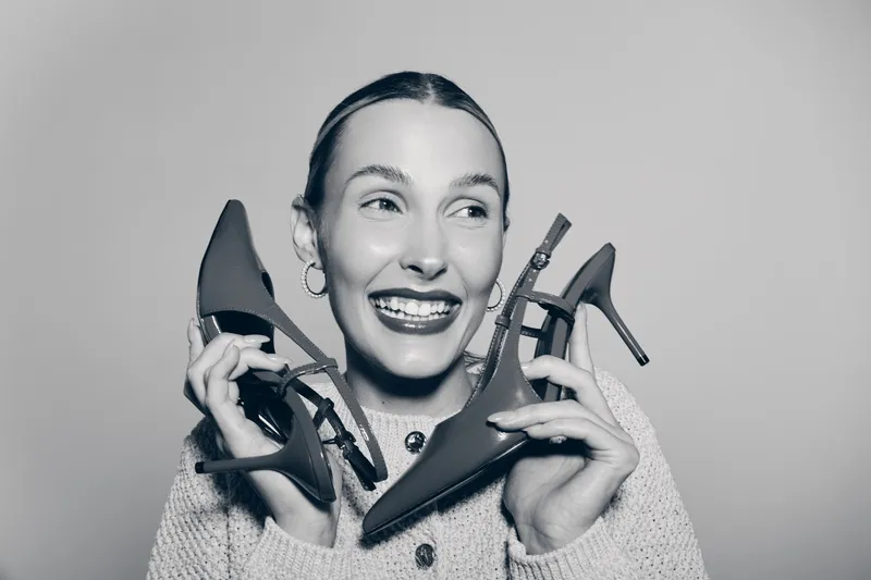 Smiling white woman with fair skin holds two high heel shoes near her face, wearing cozy sweater and hoop earrings. Concept for fashion, joy, and lifestyle with footwear accessory.