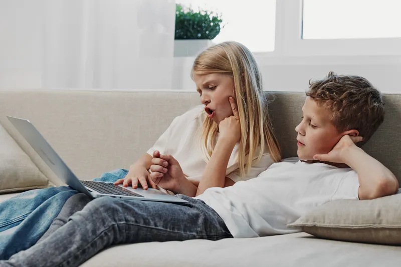 Two children, boy and girl, lie on couch using laptop together in bright room. They show curiosity and engagement, illustrating digital learning and sibling bonding at home.