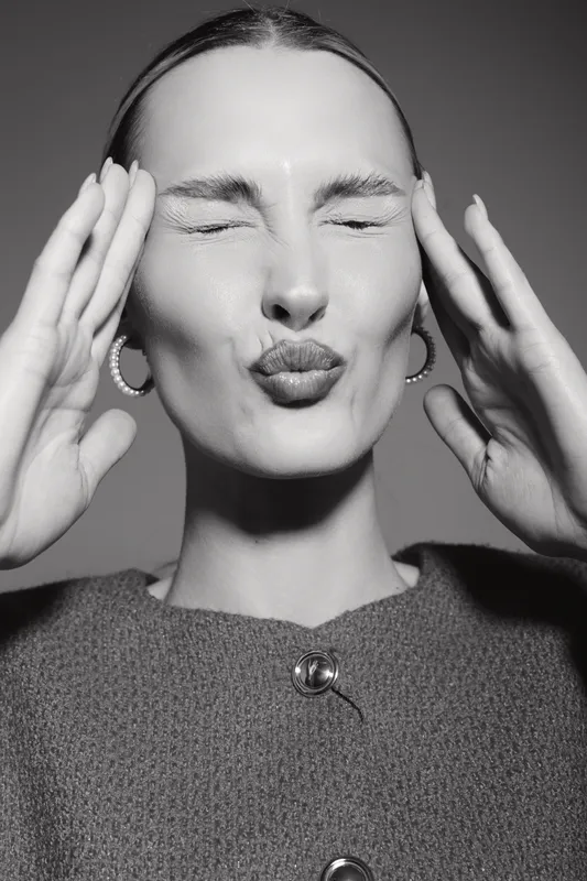 Young Caucasian woman with light skin making a playful kiss face, eyes closed, hands near head, wearing earrings and textured buttoned top in black and white portrait.