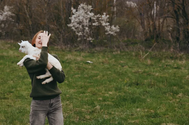 Smiling woman outdoors holding a playful goat kid, green meadow and blossoming trees behind. A warm lifestyle moment of affection, nature, and animal bonding in natural light. Warm glow.