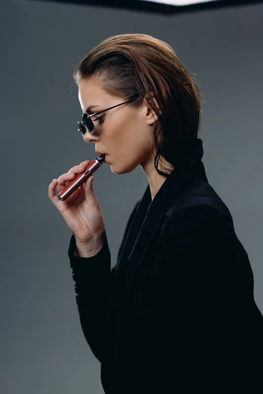 Stylish woman vaping with electronic cigarette indoors wearing sunglasses and black outfit