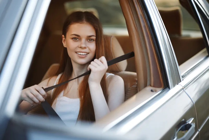 Young woman smiling and fastening seatbelt in a car, wearing a casual outfit, reflecting the concept of road safety and confidence while driving.