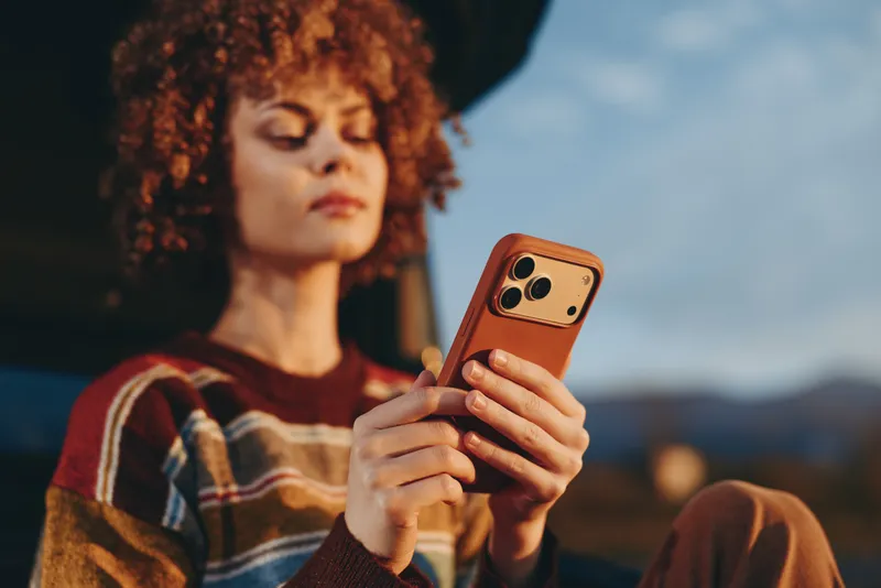 Woman smartphone portrait outdoors lifestyle curly hair tech moment, she holds phone in both hands while standing in calm evening landscape, soft focus, smiling casual leisure.