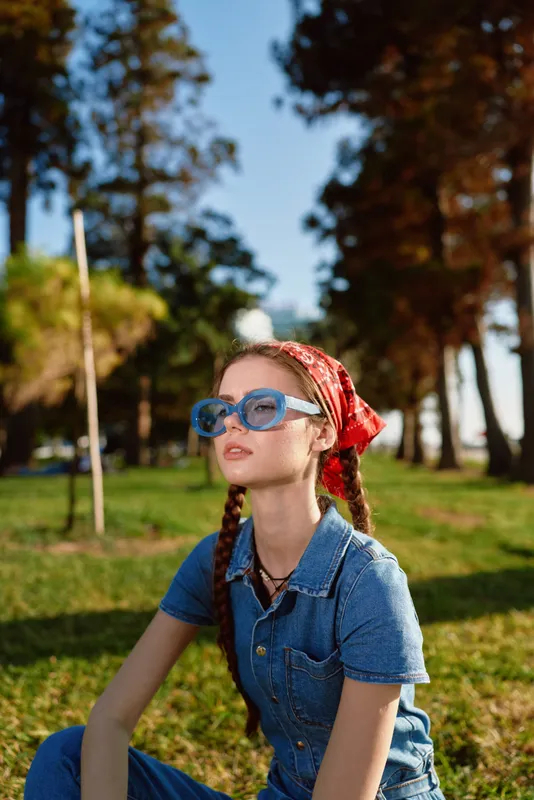 young woman wearing blue sunglasses and denim outfit sits outdoors with trees in bright sunlight and red scarf headband