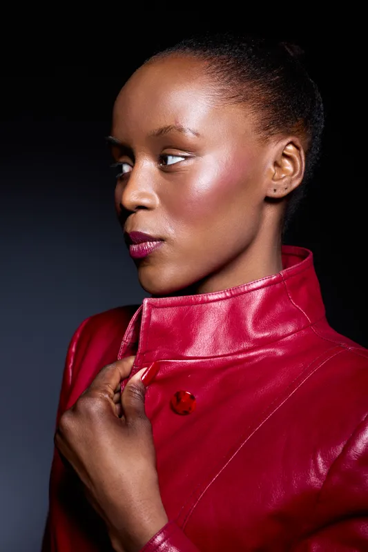 Portrait of a dark skinned African woman wearing a red leather jacket with a high collar, looking sideways. Studio lighting highlights her smooth skin and elegant facial features.