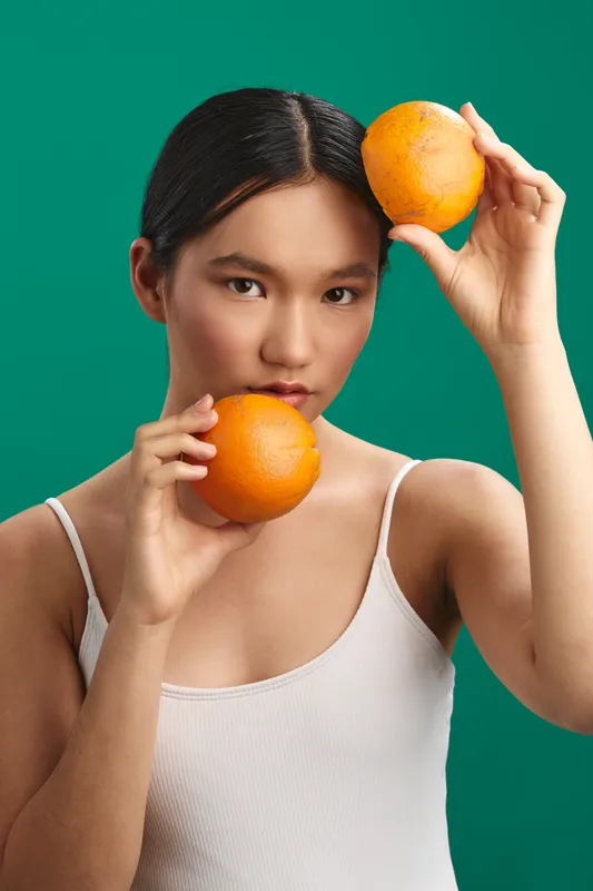 asian woman is photographed in a minimal studio setting, holding two oranges against a vivid green backdrop. she looks at the camera with a calm smile, conveying freshness, health, and natural