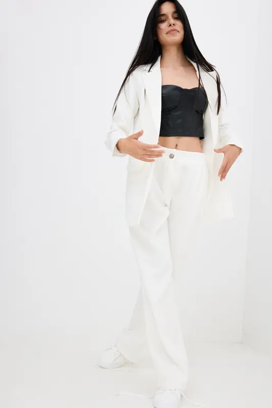 Young woman wearing white suit and black crop top standing confidently against a minimalist white background with long dark hair and casual style.