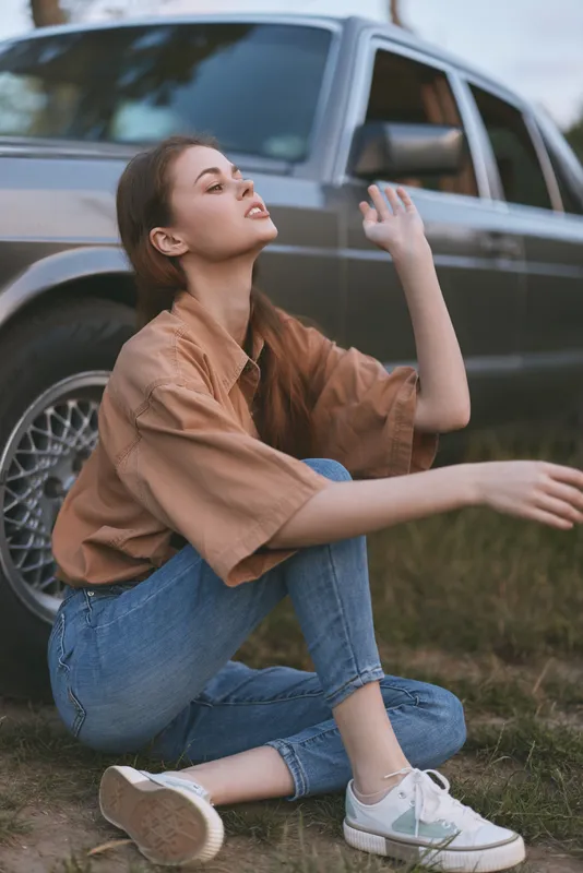 Young woman in casual attire sitting by a classic car, showcasing a relaxed vibe and natural beauty, perfect for lifestyle and fashion concepts.