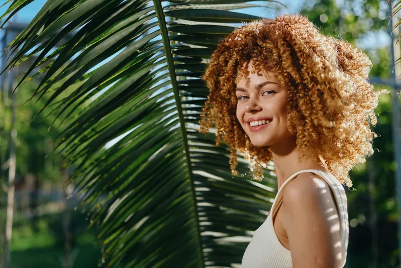 smiling woman with curly hair stands near large green palm leaf outdoors in sunny garden, wearing white sleeveless top, natural light and cheerful expression, warm atmosphere