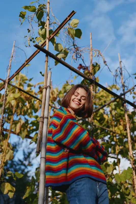 Young woman in a rainbow sweater smiling and leaning against a bamboo garden trellis with climbing vines, early twenties model in denim jeans, joyful relaxed outdoor lifestyle portrait.