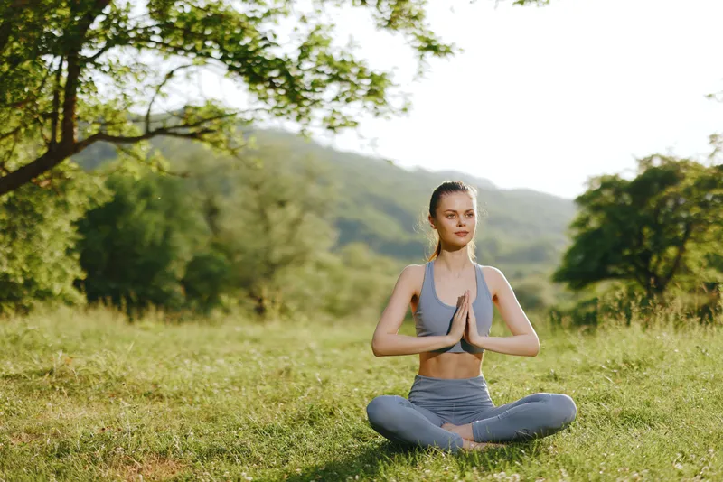 Woman practicing yoga outdoors in a serene green landscape, showcasing tranquility and wellness, dressed in athletic wear with a peaceful expression