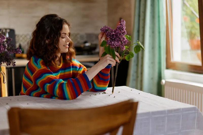 Cheerful woman indoors, sitting at a dining table, holding a lilac bouquet, colorful striped sweater, soft natural light, cozy home mood, candid lifestyle moment, bright backdrop, cozy