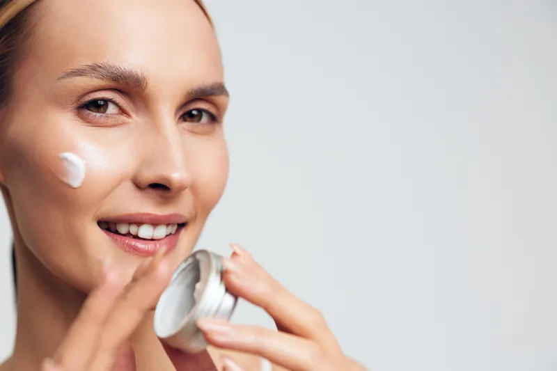 Young woman with clear skin applies white face cream on her cheek while smiling gently against a light background in a skincare and beauty concept.