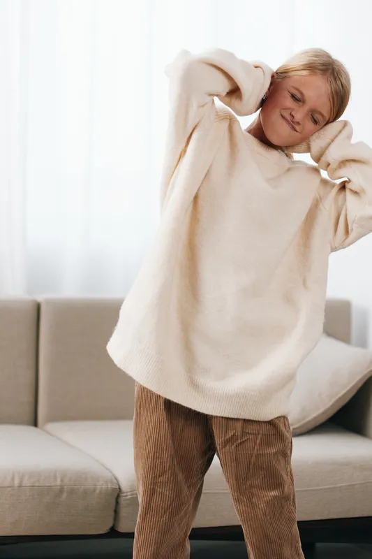 Young girl stretches in a cozy beige sweater at home, with soft natural light and a relaxed mood, casual fashion, comfortable vibe, playful pose, corduroy pants, warm atmosphere.