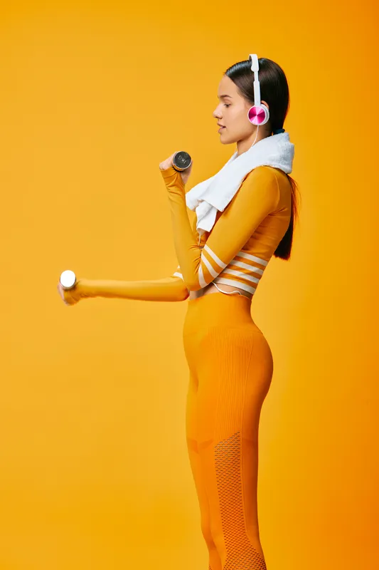 Female athlete in bright orange athletic wear with headphones and a towel around her neck, lifting a dumbbell against a vibrant orange backdrop for a vivid fitness workout