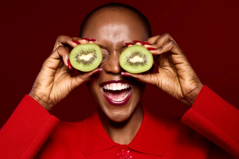 Smiling African woman with dark skin holds two kiwi halves in front of her eyes on red background. Concept of healthy lifestyle, fresh fruit, joy, and natural beauty close-up portrait.