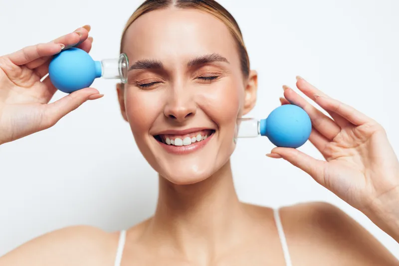 Smiling woman with closed eyes holding blue suction cups on her face. Beauty treatment concept with natural skin and relaxed expression on white background.