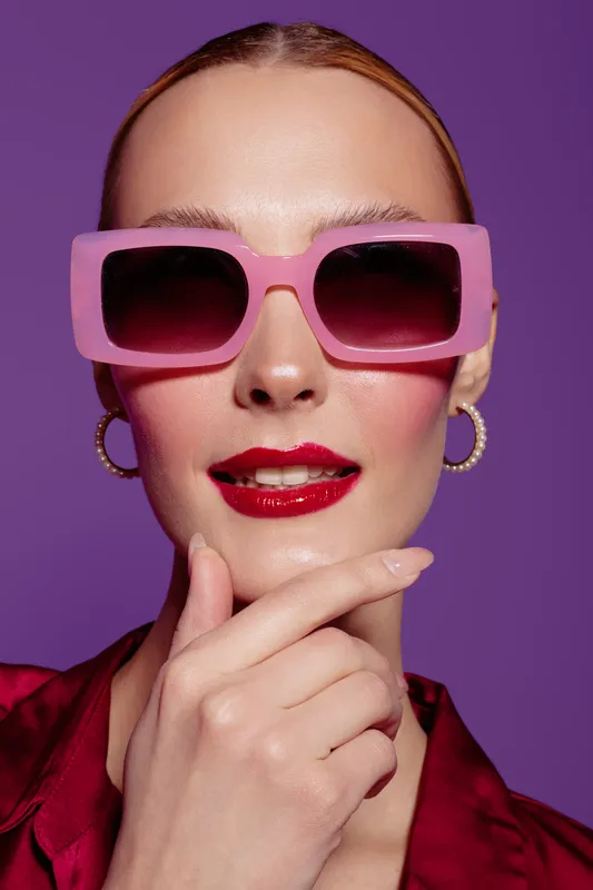 Close-up of confident woman wearing large pink sunglasses and red lipstick posing with hand near chin on purple background. Concept of fashion, style, beauty, and modern accessories.