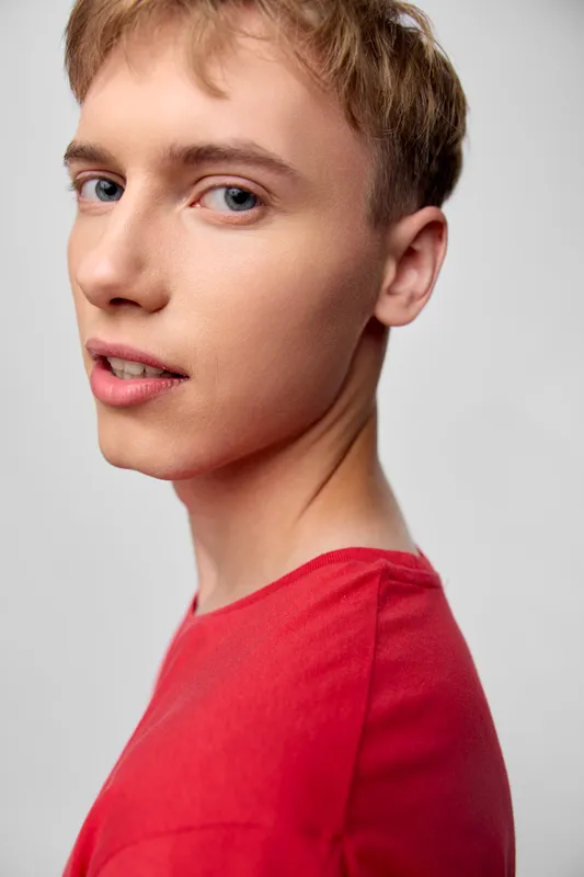 Close up of a young man in a red shirt, facing slightly to the side with a calm expression, captured in a bright studio setting for contemporary fashion and lifestyle campaigns, focusing on clean