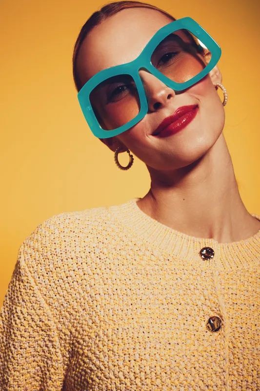 Stylish woman wearing large blue sunglasses and red lipstick smiles confidently against yellow background. Fashion concept with bright colors and modern accessories.