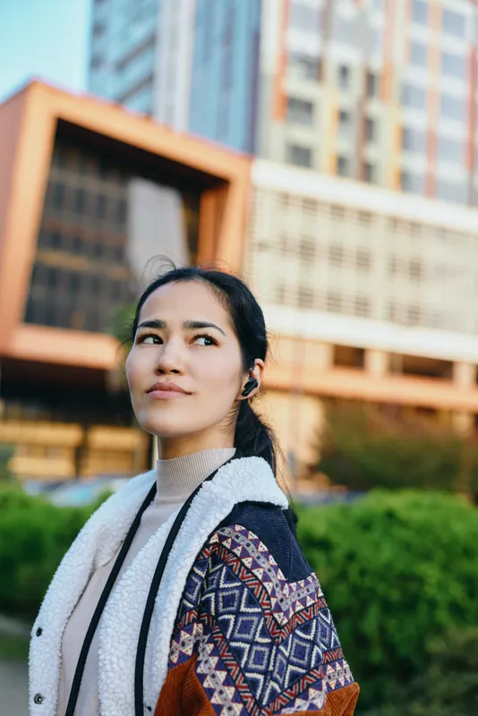 Woman in a layered coat and scarf stands outdoors in an urban setting with modern buildings behind, a calm thoughtful gaze and gentle breeze shaping her hair