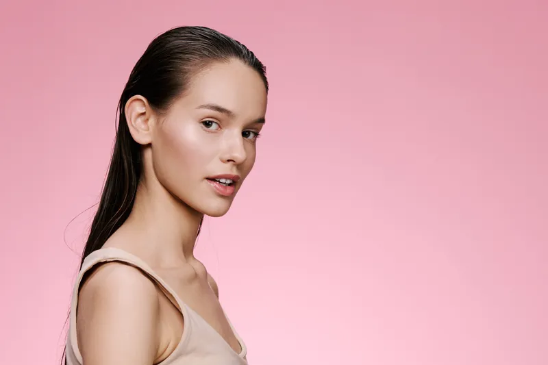 Stylish portrait of a young woman with long hair against a soft pink background, showcasing a natural look, expressing confidence and beauty in minimalist fashion