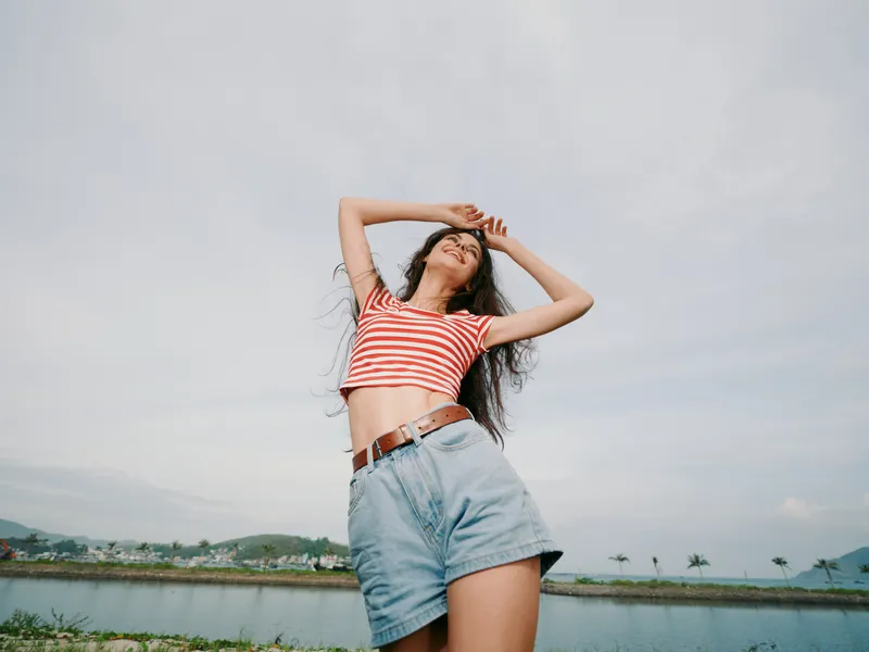 Young woman poses confidently outdoors by a calm lake, wearing a striped cropped top and denim shorts, arms raised, wind in her hair, bright sky above.