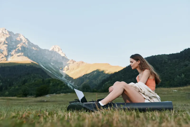 A focused professional woman sits on a mat in a grassy meadow with a laptop, surrounded by mountains. She engages with her work, radiating calm and productivity.