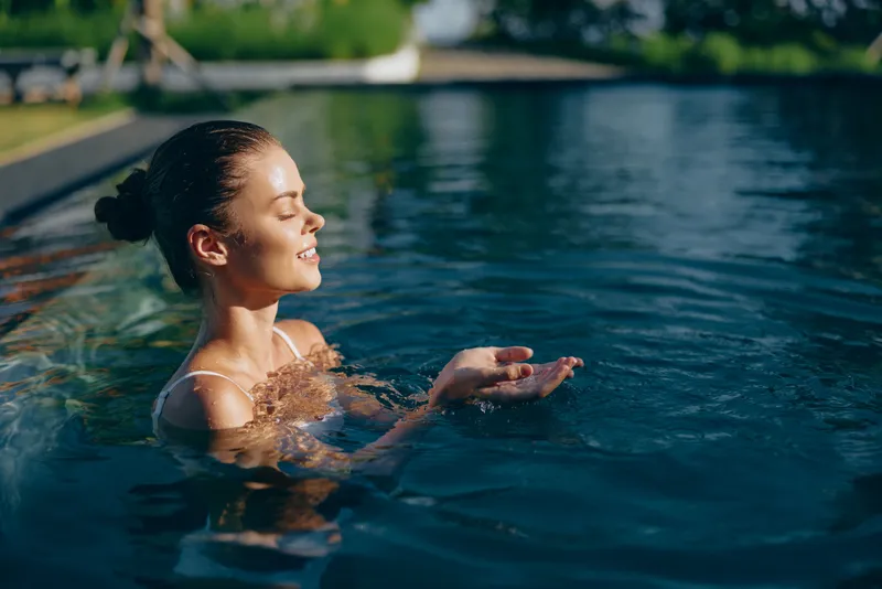 Smiling woman relaxing in a sunny pool, enjoying warm light and calm water. Lifestyle scene focused on wellness, leisure, and self care, with bright outdoor atmosphere.