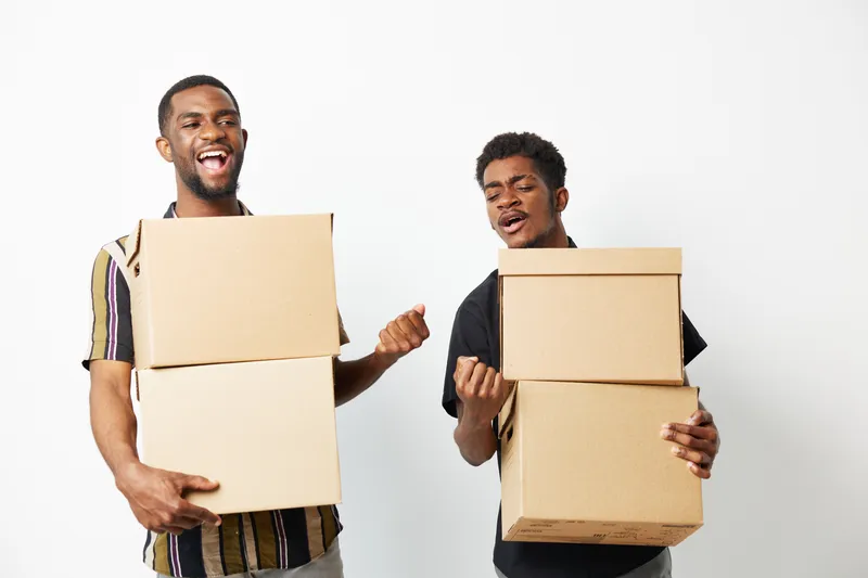 Two black men joyfully carrying boxes, showcasing teamwork and enthusiasm. Their expressions highlight a sense of camaraderie in a bright, minimalist setting.