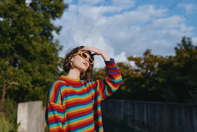 Young woman in a colorful striped sweater and yellow sunglasses posing outdoors, short hair, hand on forehead, relaxed playful expression, casual outfit for lifestyle fashion.