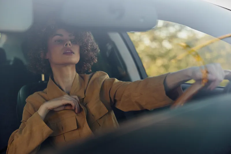 A young woman in a brown jacket drives a car with a calm, focused expression, embodying a retro film mood and a sense of independence and purposeful momentum.