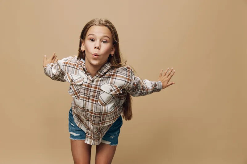 Girl in plaid shirt and denim shorts posing with arms outstretched against beige background. Portrait of playful child with long hair and casual clothing indoors.