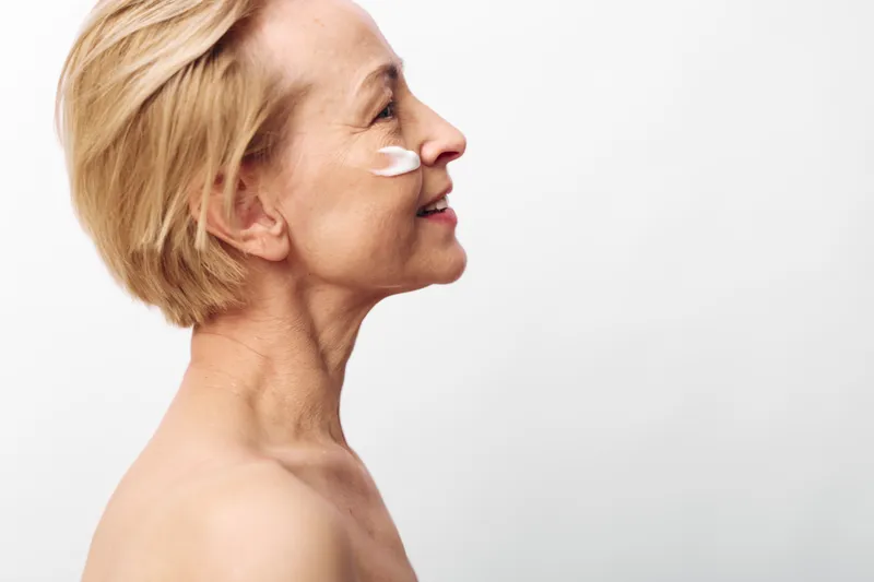 Mature woman with short blonde hair applies white cream on cheek. Profile view on white background. Concept of skincare, aging skin care, wellness, self-care, beauty routine, natural beauty.