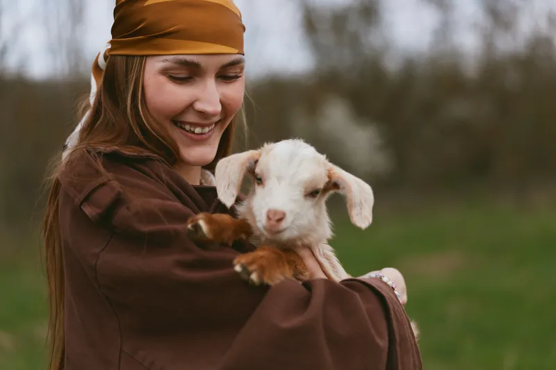 Lifestyle portrait of a smiling girl holding a gentle goat kid outdoors, brown cloak and scarf, warm light in a rustic field, showing affection and companionship in a pastoral moment