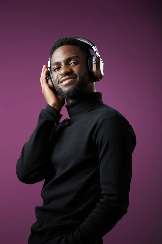 Confident young man enjoying music with headphones, wearing a black turtleneck against a purple background, smiling with a relaxed expression