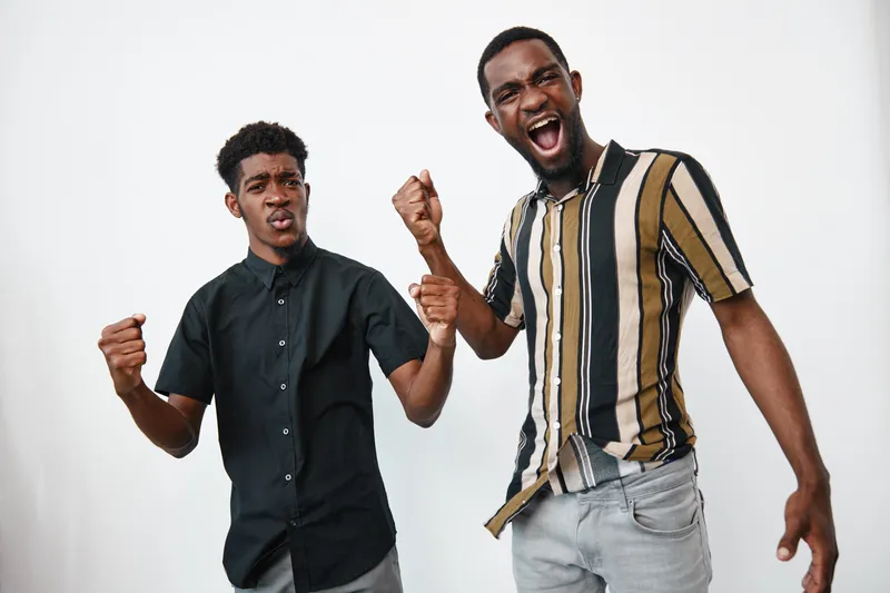 Two young black men with dark skin celebrating and showing excitement together in casual clothes on white background. Friendship, happiness and energy in studio portrait.