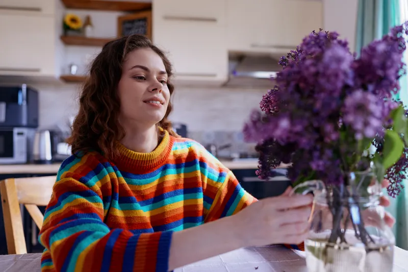 Joyful woman indoors at home, wearing a colorful striped sweater, reaching toward a vase of purple flowers, warm natural light, cozy kitchen backdrop, soft fabrics, candid lifestyle