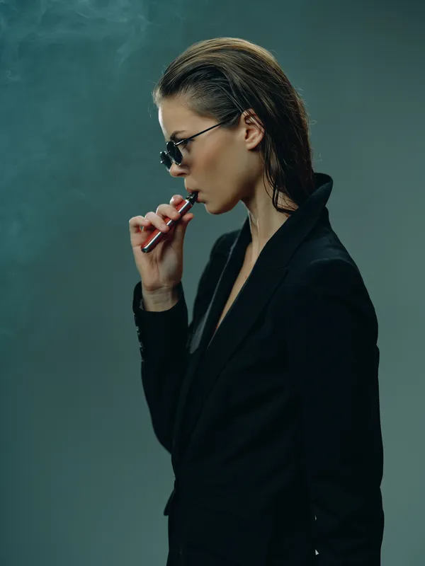 Woman vaping with electronic cigarette in black outfit wearing sunglasses with smoky background