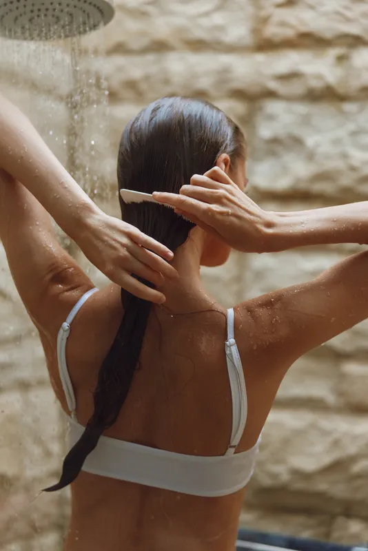 Woman combing wet hair under outdoor shower with stone wall background, relaxed mood, natural light, summer wellness, healthy hair care routine.