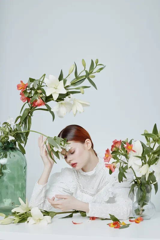 Elegant woman in white attire surrounded by colorful flowers on a minimalistic background, portraying serenity and natural beauty in a fashion photo shoot
