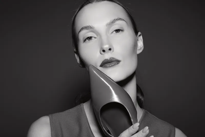 Caucasian woman with pale skin holds a shiny high heel shoe near her face. Studio portrait in black and white with soft lighting. Concept for fashion and elegance.