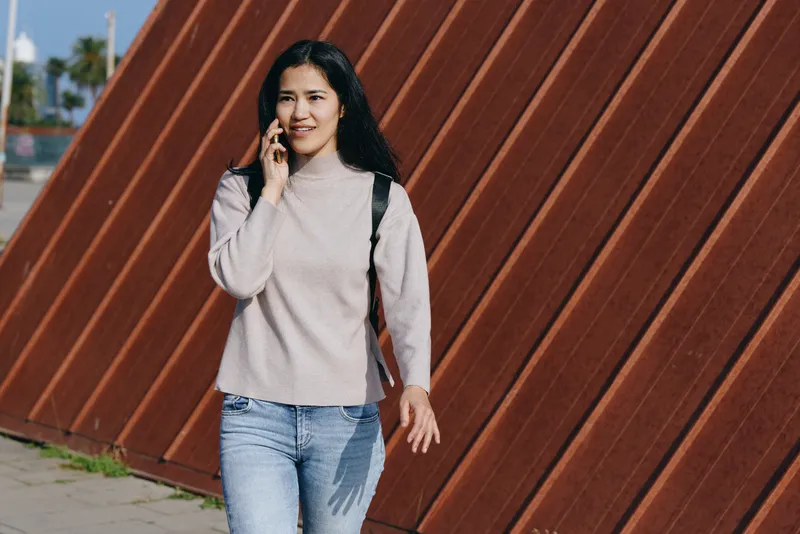 Woman talking on phone while walking outdoors beside a rust brown diagonal wall, wearing a light gray sweater and jeans, casual urban moment captured in daylight