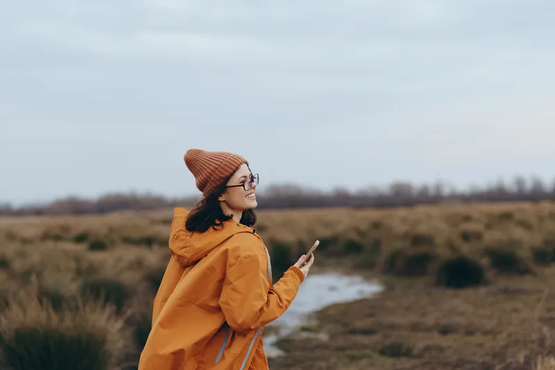 Woman lifestyle outdoors autumn, orange coat and knit hat, standing in a rural field, wind blows hair, calm nature mood, cloudy sky, wide grassy landscape, distant horizon, soft light