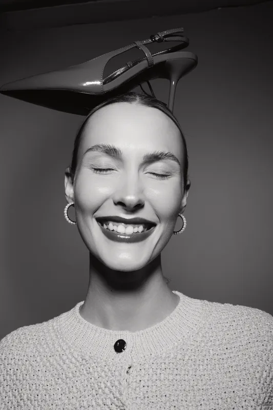 Smiling white woman with light skin and closed eyes balances high heel shoe on head in studio portrait. Concept of fashion, joy, and creativity.